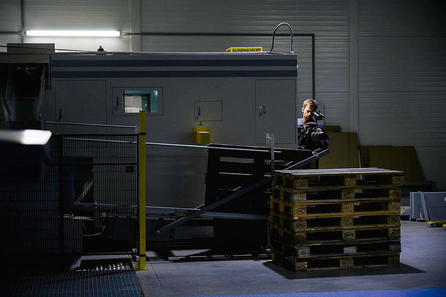 Ein Mann im Halbdunkel hinter einem Stapel Paletten sieht durch eine Kamera