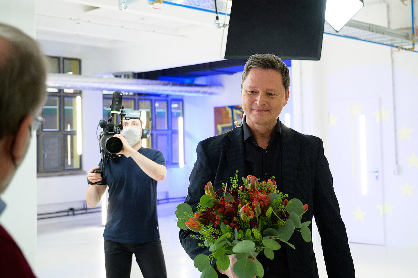 Zum Geburtstag werden Andreas Fritsch durch Volker Kurz ein Strauß Blumen überreicht. Eine Kamera filmt das geschehen.