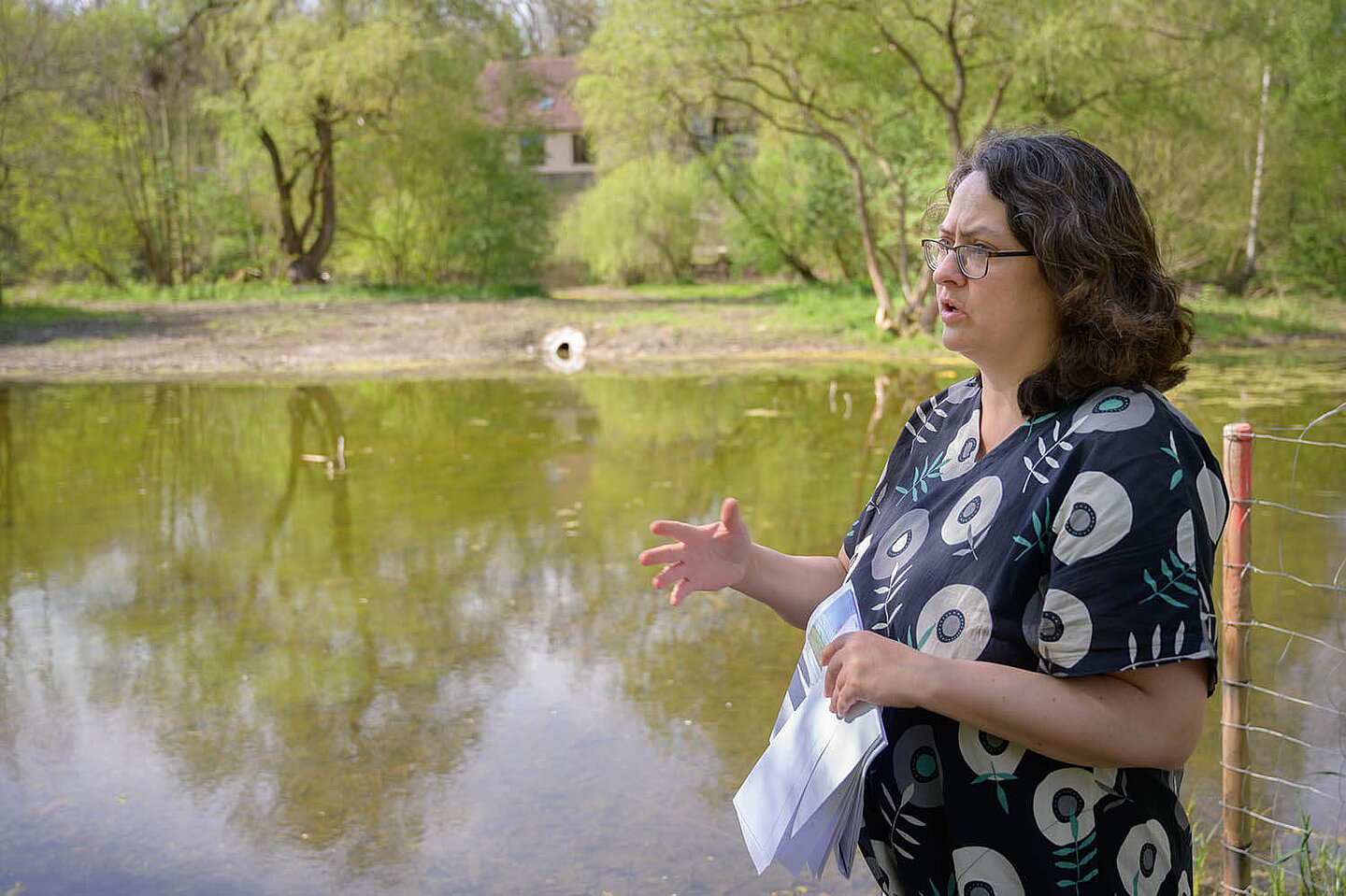A woman standing in front of a pond explains something to a person who is not visible while gesturing