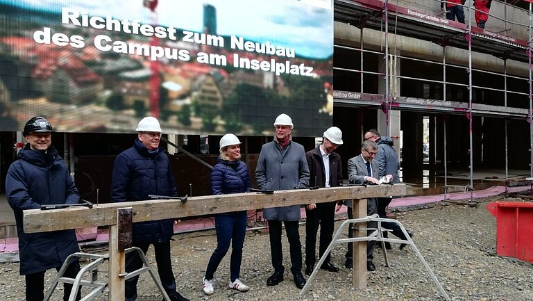 6 Personen mit Schutzhelm stehen vor einer Videoprojektion mit der Aufschrift: Richtfest zum Neubau des Campus am Inselplatz, dahinter ein Gebäude im Rohbau