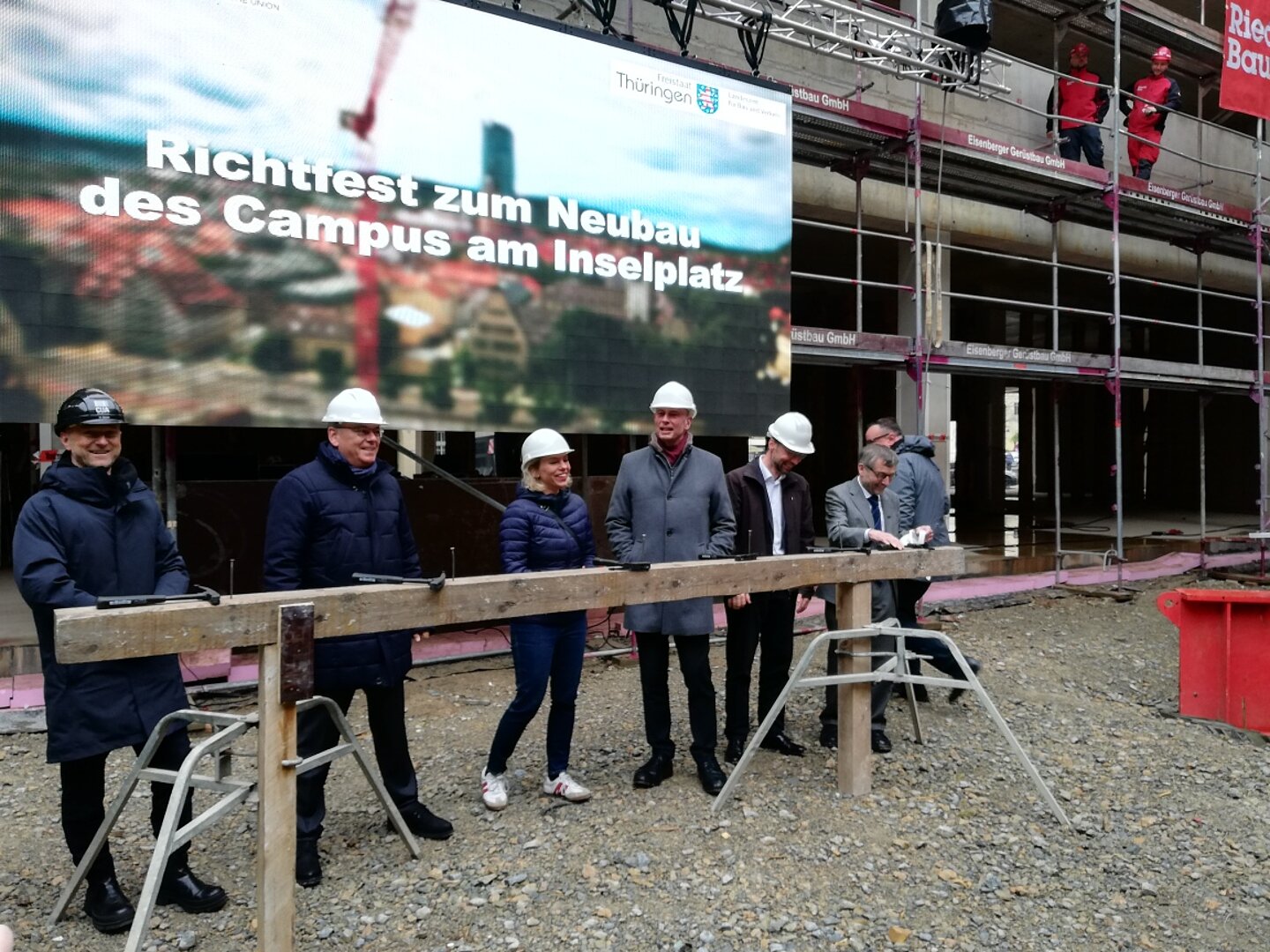 6 Personen mit Schutzhelm stehen vor einer Videoprojektion mit der Aufschrift: Richtfest zum Neubau des Campus am Inselplatz, dahinter ein Gebäude im Rohbau