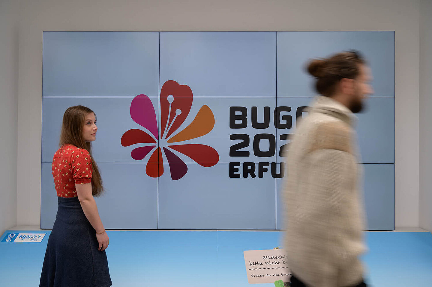 Ein Mann und eine Frau welche vor einem Bildschirm stehen, auf dem das Logo der BUGA 2021 abgebildet ist