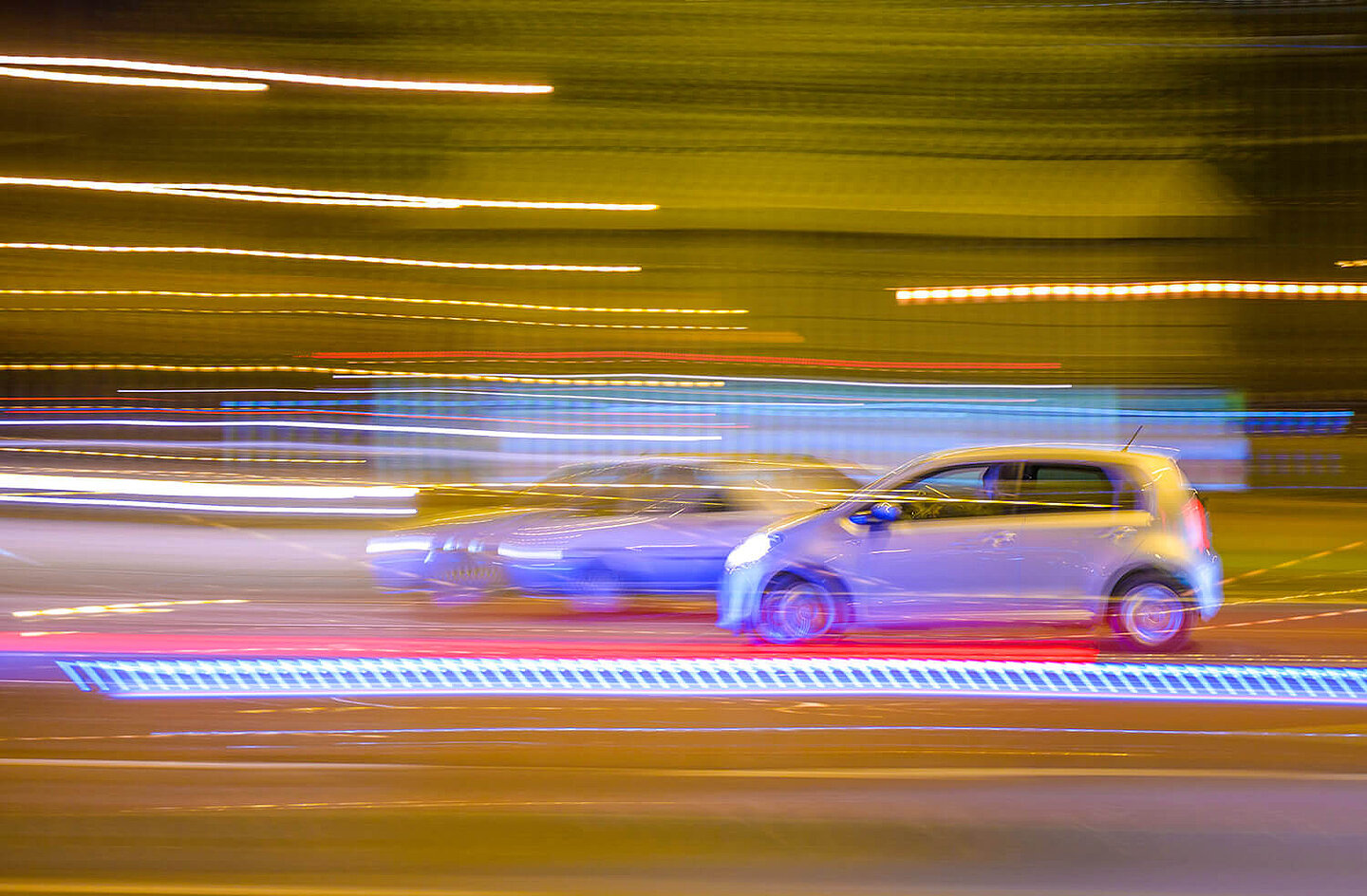 Zwei bei Nacht vorbeifahrende Autos. Autos und Lichter sind durch die Geschwindigkeit nur verschwommen zu sehen.