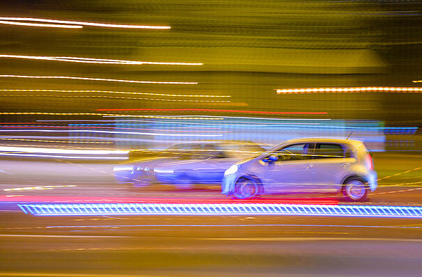 Zwei bei Nacht vorbeifahrende Autos. Autos und Lichter sind durch die Geschwindigkeit nur verschwommen zu sehen.