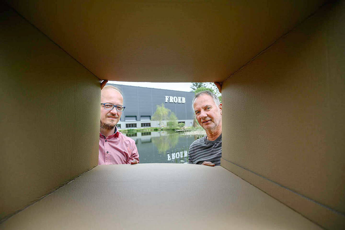 Zwei Männer sehen durch einen Papkarton in die Kamera. Hinter ihnen befindet sich ein Haus der Froeb-Verpackungen GmbH.