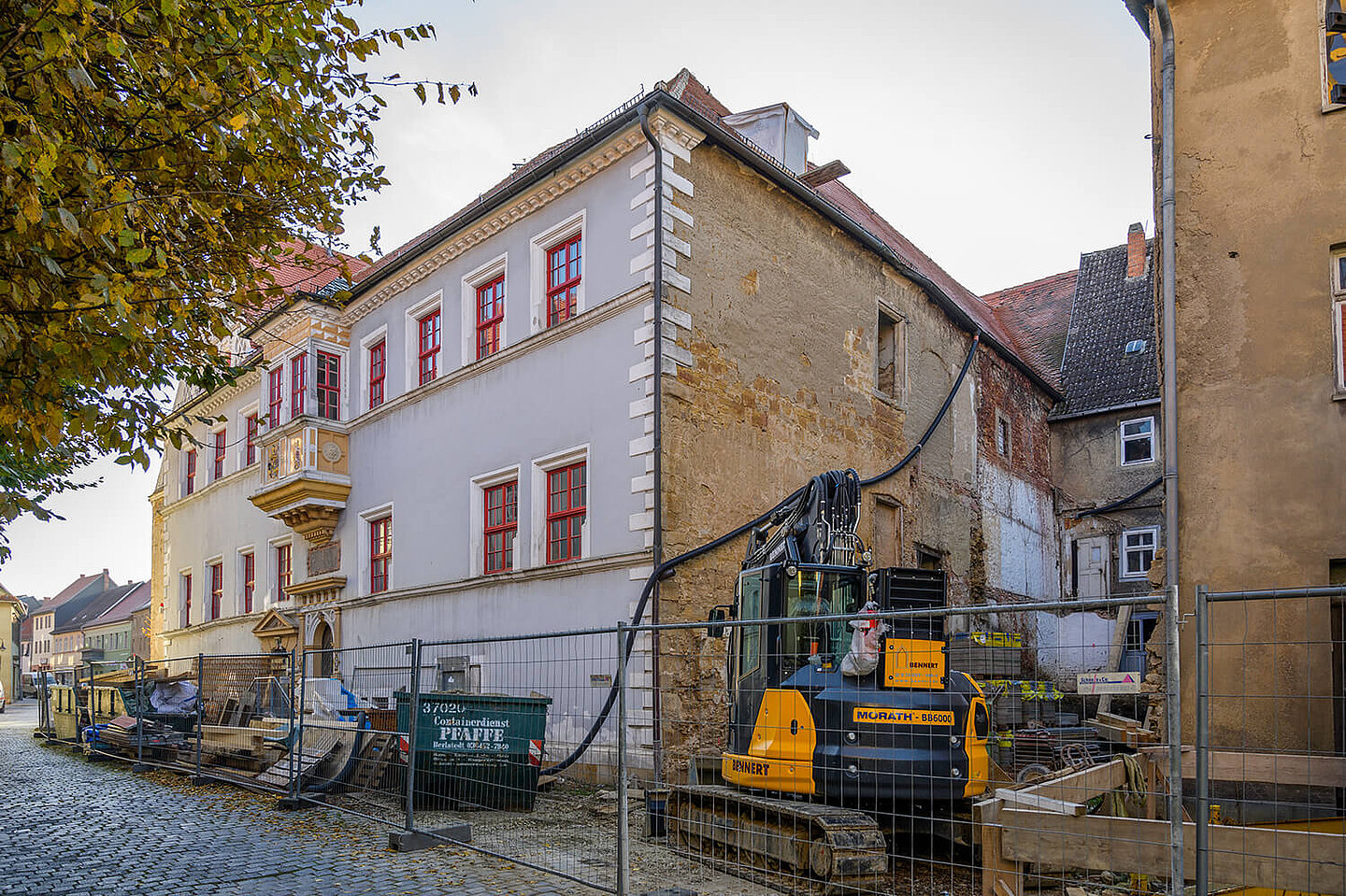 Baustelle am Rathaus. Ein Bagger und mehrere Schuttcontainer sind mit einem Bauzaun umschlossen.