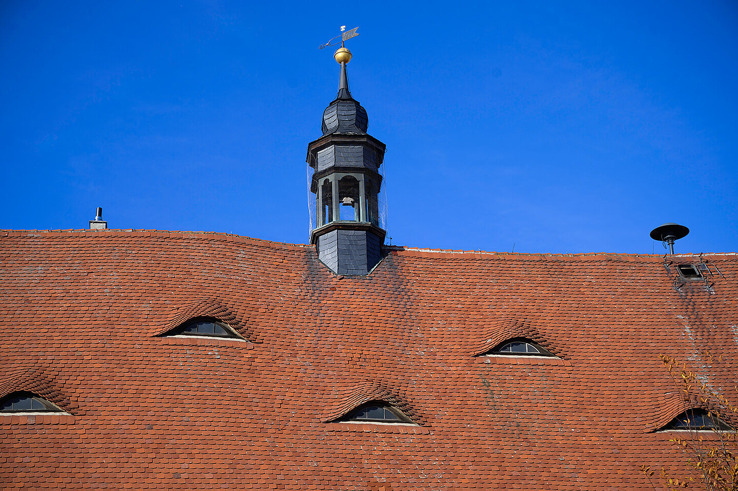 Glockenturm vom Buttstädter Rathaus. Dieser sitzt auf einem roten Dach mit kleinen Fenstern. In einer Ecke ist eine Alarmsirene montiert.