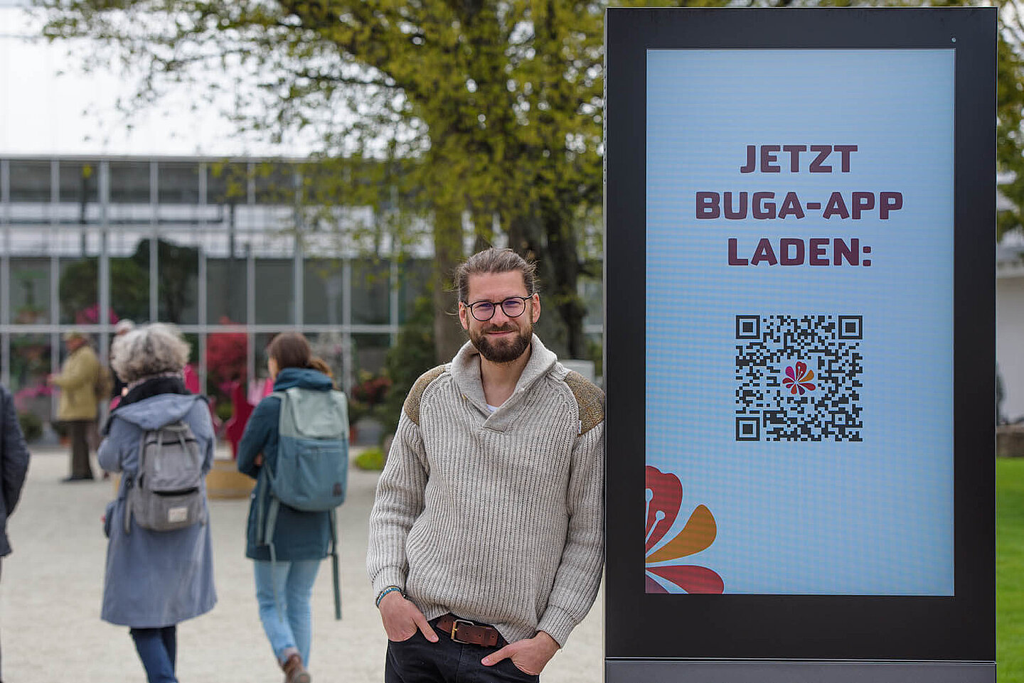 Ein Mann, welcher sich an eine Werbetafel der BUGA 2021 lehnt. Im Hintergrund befinden sich gehende Menschen.